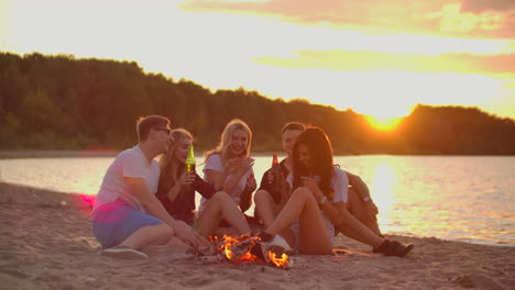 Five-young-people-are-sitting-around-bonfire-on-the-sand-beach.-They-are-talking-to-each-other-and-drinking-beer-at-sunset-and-enjoying-the-summer-evening-on-the-lake-coast.